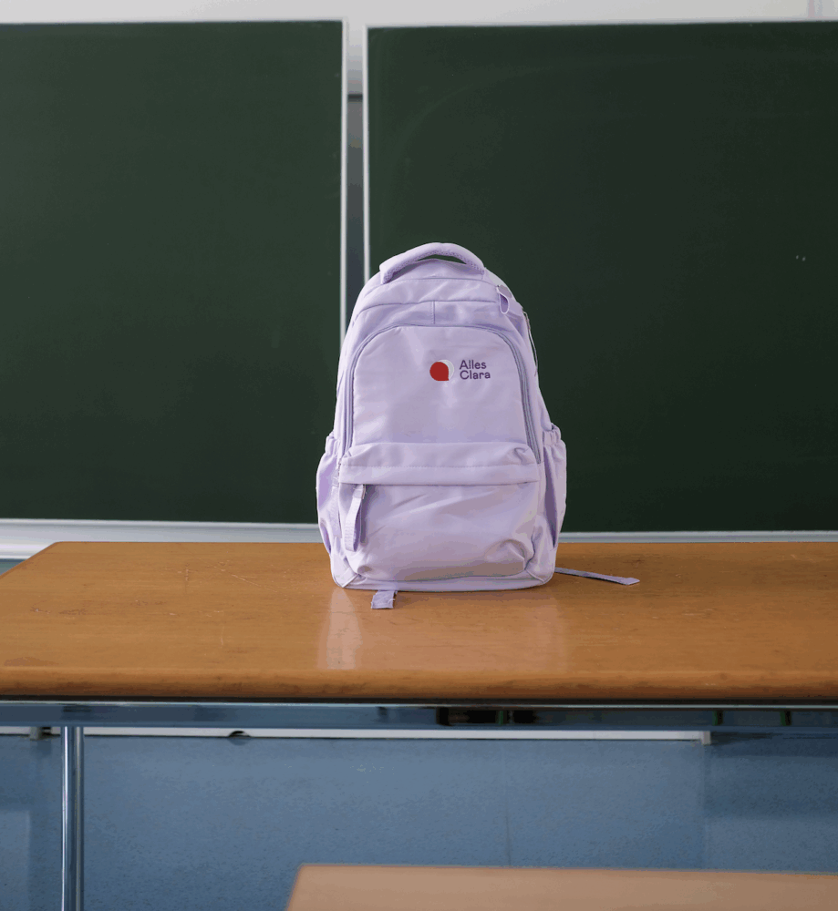 Ein lila Rucksack mit dem „Alles Clara“-Logo auf einem Tisch vor einer Tafel