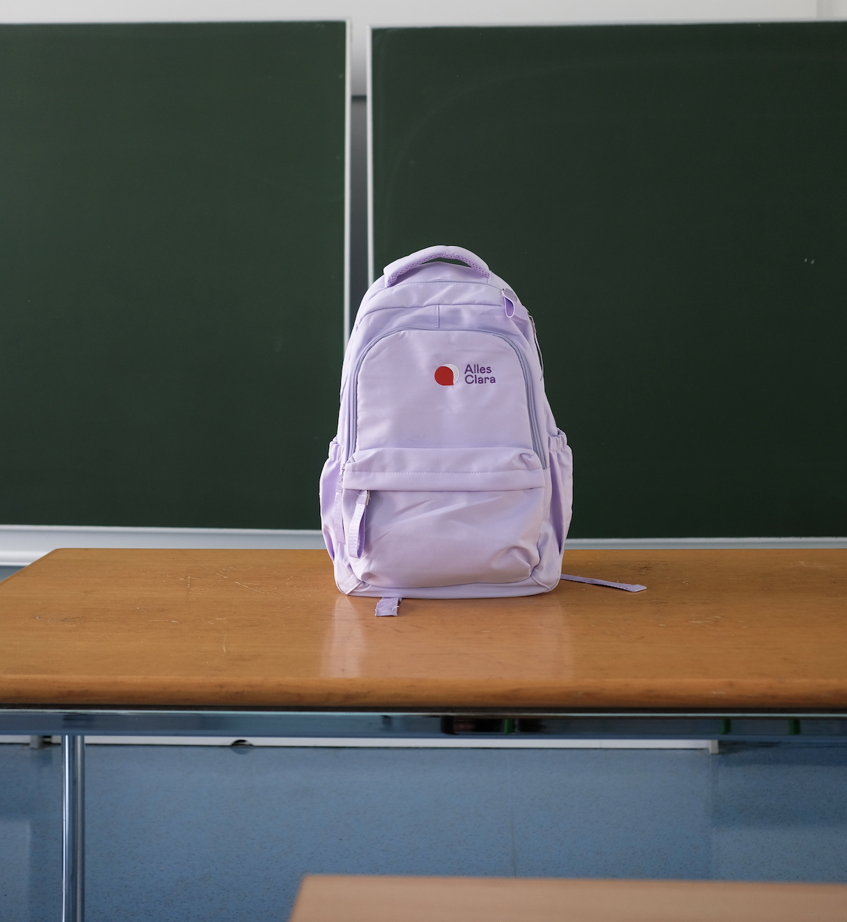 Purple backpack with the Alles Clara logo on a table in front of a blackboard